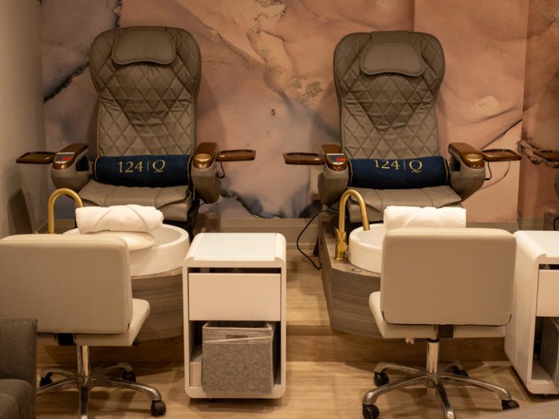 A cozy nail salon setup featuring two plush pedicure chairs with armrests. Each chair faces a foot basin with rolled white towels on the edge. A small white table with a storage basket is placed between the chairs. The wall has abstract artwork. 124 on Queen Hotel and Spa | Hotel & Spa Niagara-on-the-lake | 1 (855) 988-4552
