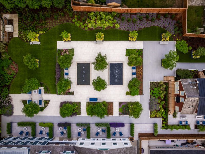 Aerial view of a landscaped garden with symmetrical pathways, trees, and shrubs. Central area features square paved sections with fountains and seating, surrounded by green lawns and flowerbeds. Buildings and wooden fences border the garden. 124 on Queen Hotel and Spa | Hotel & Spa Niagara-on-the-lake | 1 (855) 988-4552