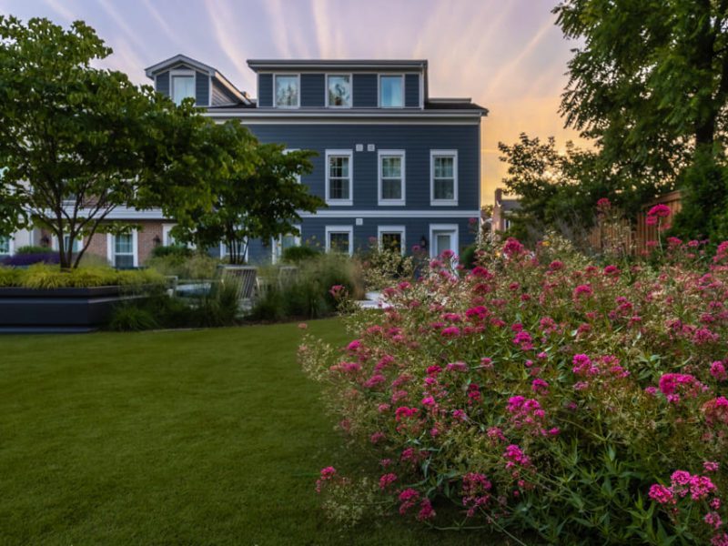 A picturesque garden with vibrant pink flowers and lush green lawns in the foreground. A large, grey-blue, three-story house with white accents is in the background, framed by leafy trees. The sky is tinged with warm hues of sunset. 124 on Queen Hotel and Spa | Hotel & Spa Niagara-on-the-lake | 1 (855) 988-4552