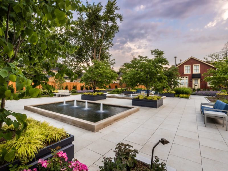 A landscaped garden with paved walkways features several rectangular water fountains surrounded by lush green plants. There are benches and blue cushioned chairs. A house is visible in the background under a partly cloudy sky. 124 on Queen Hotel and Spa | Hotel & Spa Niagara-on-the-lake | 1 (855) 988-4552