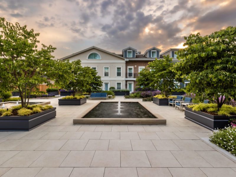 Rooftop garden with large rectangular water feature, surrounded by lush trees and planters. White tiles cover the ground. Three-story building with gabled roof in the background under a dramatic, cloud-filled sky. 124 on Queen Hotel and Spa | Hotel & Spa Niagara-on-the-lake | 1 (855) 988-4552