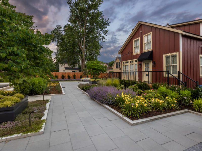 A landscaped yard with a pathway, flowerbeds, and a red house with cream trim under a cloudy sky. Tall trees and shrubs are in the background, with colorful flowers and lavender lining the path. 124 on Queen Hotel and Spa | Hotel & Spa Niagara-on-the-lake | 1 (855) 988-4552