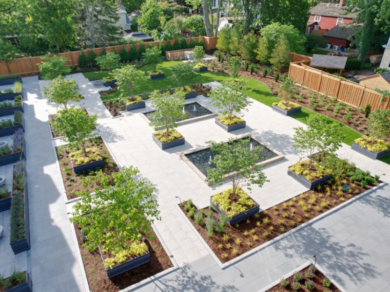 A symmetrical garden with neatly arranged planters and small trees on a spacious, paved courtyard. Surrounded by a wooden fence and trees, the area is well-maintained, with pathways and evenly distributed greenery creating a serene, orderly appearance. 124 on Queen Hotel and Spa | Hotel & Spa Niagara-on-the-lake | 1 (855) 988-4552