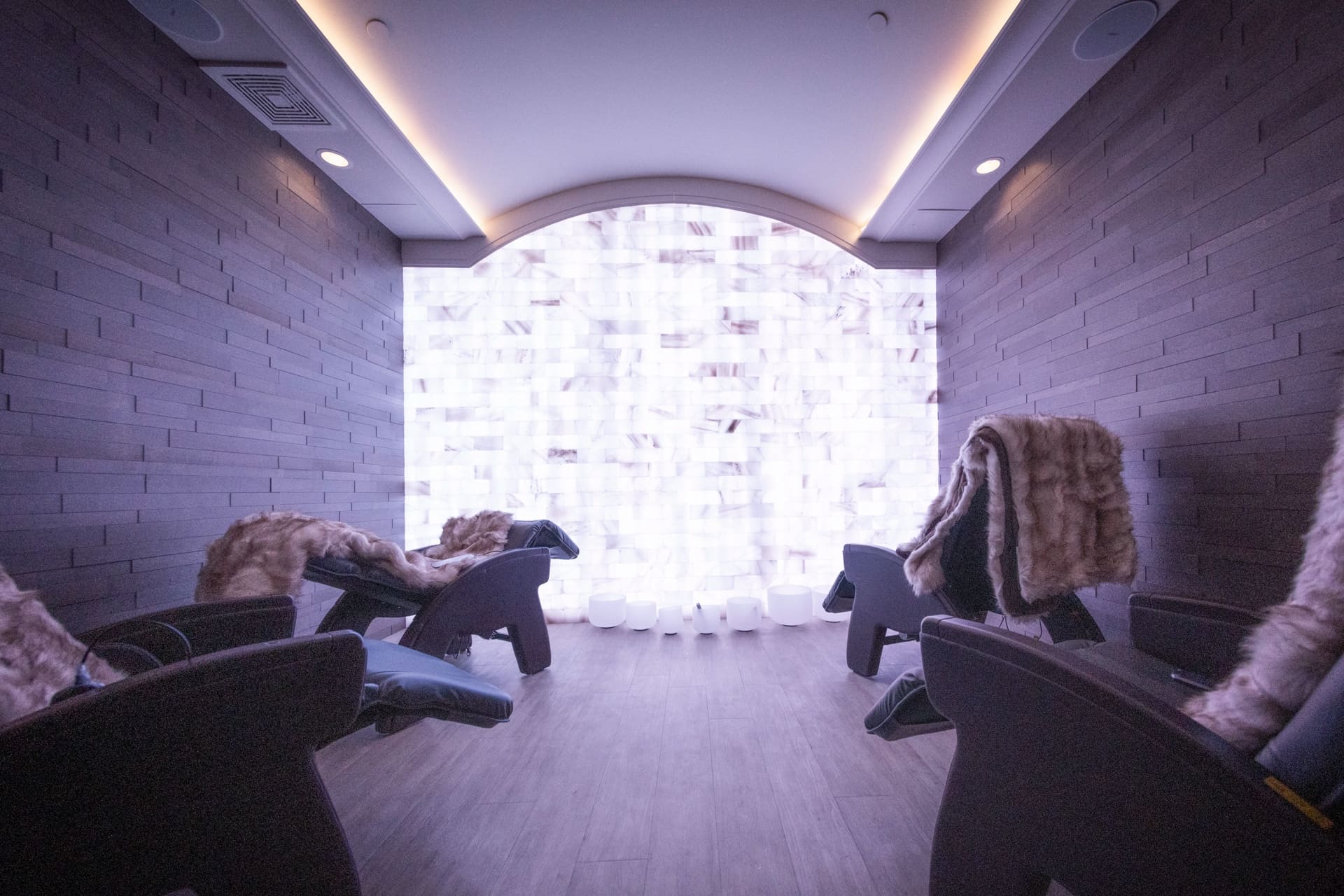 A serene spa room with four reclining chairs draped in fur throws. The wood-paneled walls are dimly lit, and a glowing wall with a mosaic pattern creates a calming atmosphere. The soft lighting enhances the tranquil ambiance. 124 on Queen Hotel and Spa | Hotel & Spa Niagara-on-the-lake | 1 (855) 988-4552