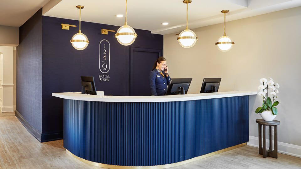A hotel reception area with a receptionist at a navy curved desk, equipped with two computers. Behind, theres a dark blue wall with a sign reading 124 Hotel & Spa. Three globe lights hang above, and a small table with white orchids sits to the side. 124 on Queen Hotel and Spa | Hotel & Spa Niagara-on-the-lake | 1 (855) 988-4552