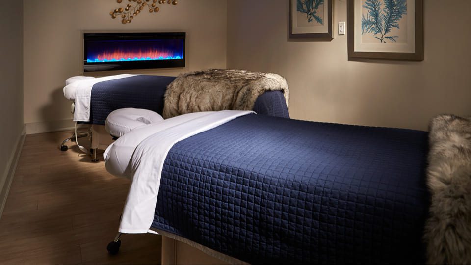 Two massage tables are adorned with dark blue covers and white sheets, each topped with a soft brown blanket. A wall-mounted electric fireplace casts a colorful glow, and framed botanical prints decorate the light-colored wall above. 124 on Queen Hotel and Spa | Hotel & Spa Niagara-on-the-lake | 1 (855) 988-4552