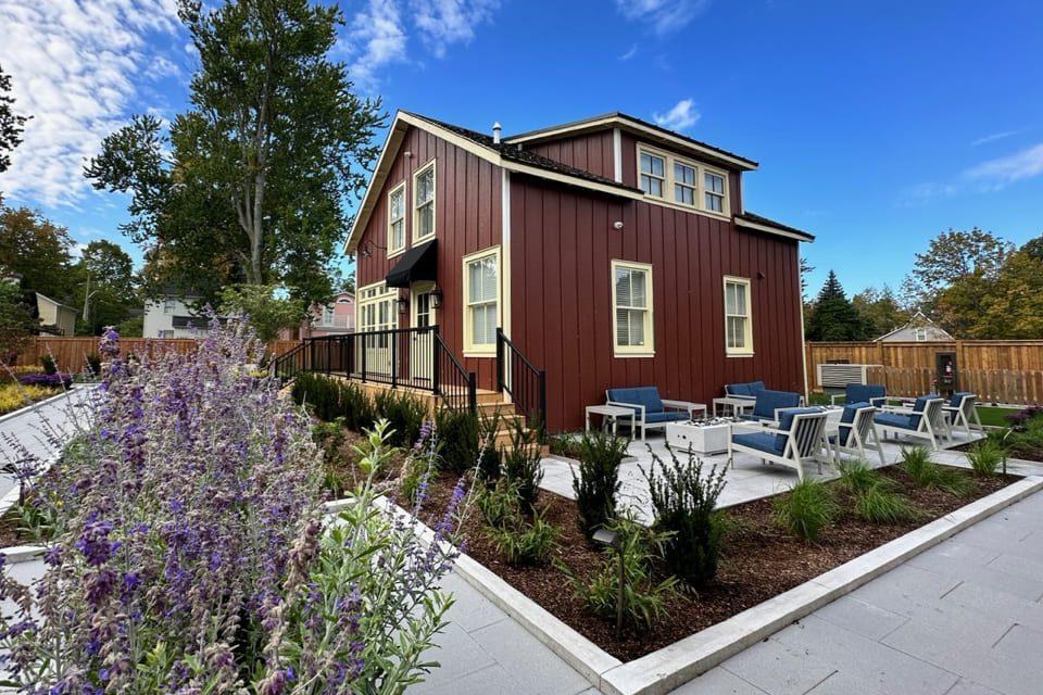 A two-story red house with white trim and a black roof is surrounded by purple flowers and landscaping. The patio features blue cushioned chairs and tables. The sky is partly cloudy with trees in the background. 124 on Queen Hotel and Spa | Hotel & Spa Niagara-on-the-lake | 1 (855) 988-4552