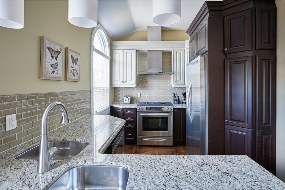 Modern kitchen with granite countertops, double sink, stainless steel appliances, and a tiled backsplash. Dark wood cabinets contrast with light walls. Two butterfly prints hang on the wall near large windows. 124 on Queen Hotel and Spa | Hotel & Spa Niagara-on-the-lake | 1 (855) 988-4552