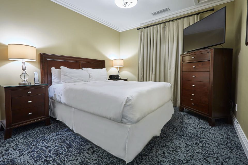 A cozy bedroom with a neatly made bed covered in white linens, flanked by two nightstands with lamps. A wooden dresser with a TV on top is against the wall, and beige curtains hang over the window. The carpet is a blue-gray pattern. 124 on Queen Hotel and Spa | Hotel & Spa Niagara-on-the-lake | 1 (855) 988-4552