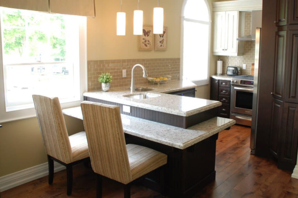 A modern kitchen features a central island with a double-tiered granite countertop and two striped upholstered chairs. Pendant lights hang above, and stainless steel appliances are visible. Light streams in through a window, illuminating the space. 124 on Queen Hotel and Spa | Hotel & Spa Niagara-on-the-lake | 1 (855) 988-4552