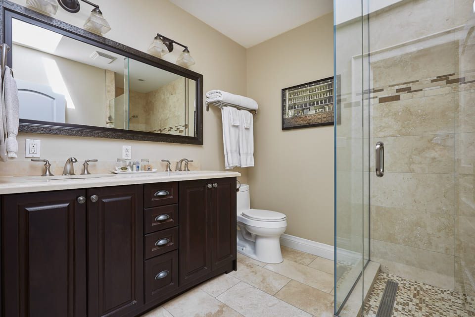 A modern bathroom with a wooden double-sink vanity, framed mirror, and wall-mounted lights. A toilet is next to a stack of towels. A glass-enclosed shower with beige tile and decorative accents is on the right. A framed picture hangs above the toilet. 124 on Queen Hotel and Spa | Hotel & Spa Niagara-on-the-lake | 1 (855) 988-4552