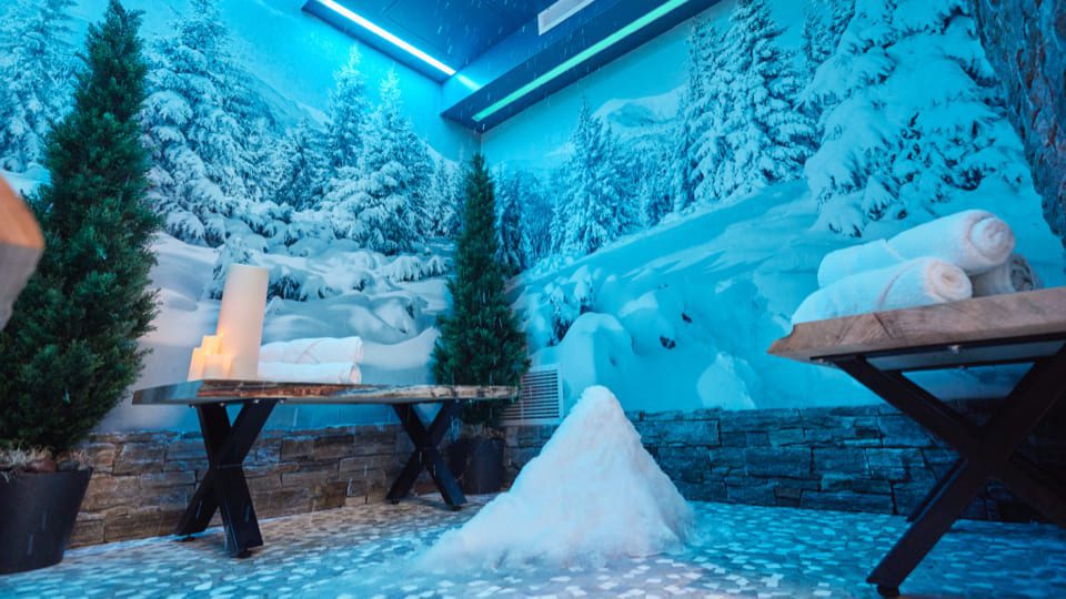 Room designed like a snowy forest with snowy walls and ceiling, snow mounds on the floor, two wooden benches with towels, and potted evergreen trees. Lit candles add warmth. 124 on Queen Hotel and Spa | Hotel & Spa Niagara-on-the-lake | 1 (855) 988-4552