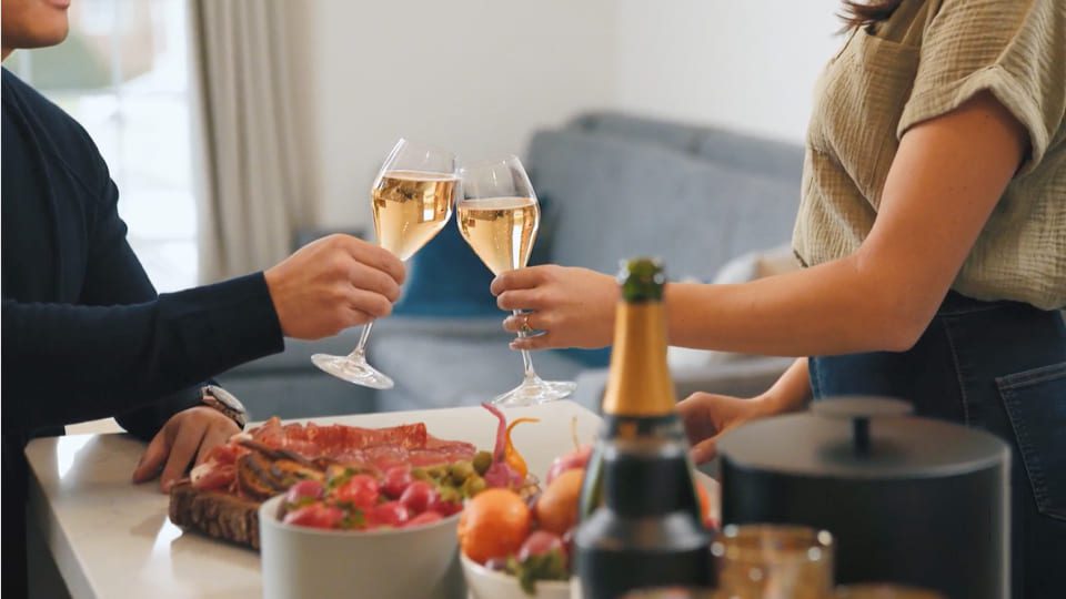 Two people clinking champagne glasses over a table with a charcuterie board, fruits, and a champagne bottle. They are in a cozy setting with soft lighting. 124 on Queen Hotel and Spa | Hotel & Spa Niagara-on-the-lake | 1 (855) 988-4552