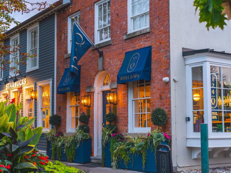 A charming street scene with a brick hotel and spa featuring blue awnings and potted plants outside. Next door is a Starbucks with a welcoming facade. Lush greenery and colorful flowers adorn the sidewalk. 124 on Queen Hotel and Spa | Hotel & Spa Niagara-on-the-lake | 1 (855) 988-4552
