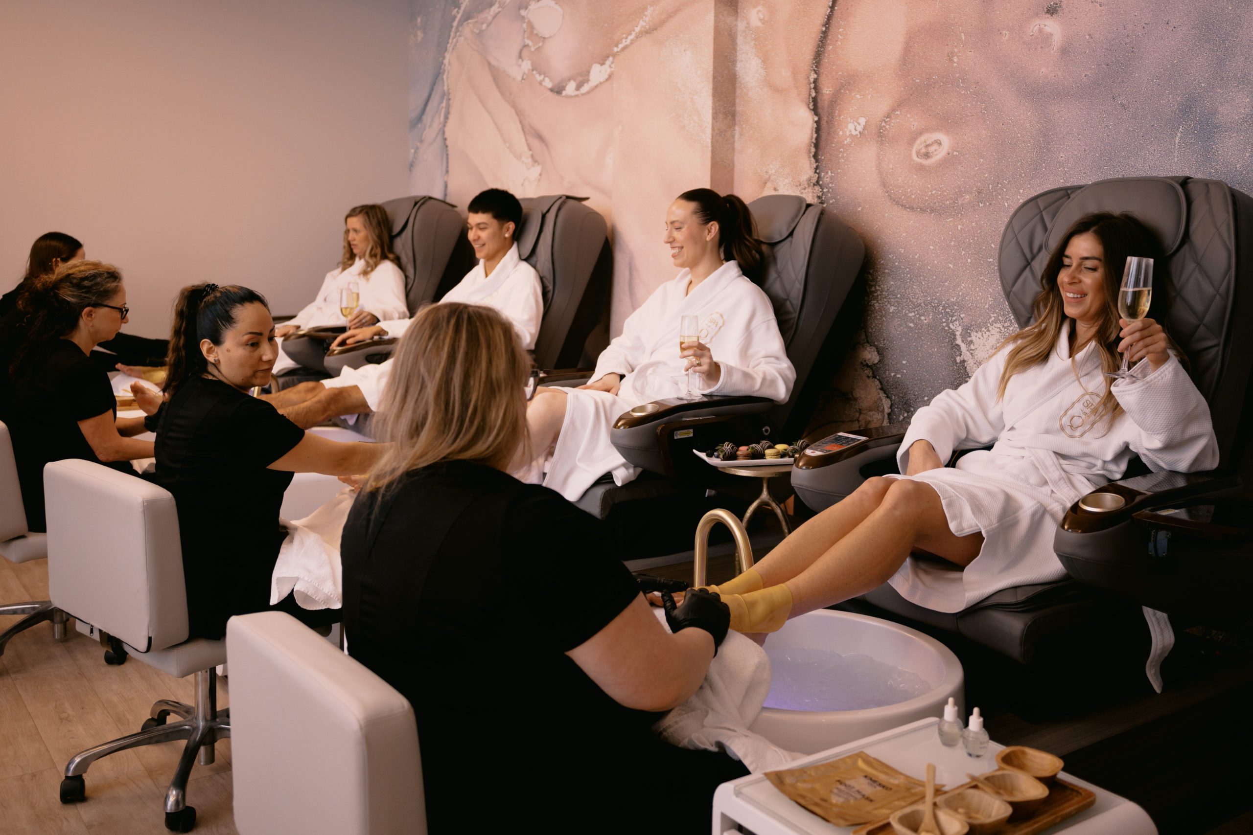 Four people sitting in massage chairs receive pedicures from technicians in a spa. They wear white robes, and some hold drinks. The atmosphere is relaxed and comfortable, with soothing decor on the walls. 124 on Queen Hotel and Spa | Hotel & Spa Niagara-on-the-lake | 1 (855) 988-4552