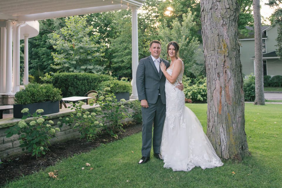 A couple in wedding attire stands by a tree in a garden. The groom is in a gray suit, and the bride is in a white gown. They are smiling and embracing, with greenery and a building in the background. 124 on Queen Hotel and Spa | Hotel & Spa Niagara-on-the-lake | 1 (855) 988-4552