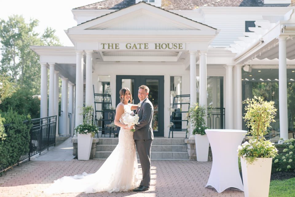 A bride and groom stand smiling in front of The Gate House. The bride holds a bouquet, and both are dressed in wedding attire. They stand on a brick path with greenery and large planters nearby. The building has tall white columns and a glass entrance. 124 on Queen Hotel and Spa | Hotel & Spa Niagara-on-the-lake | 1 (855) 988-4552