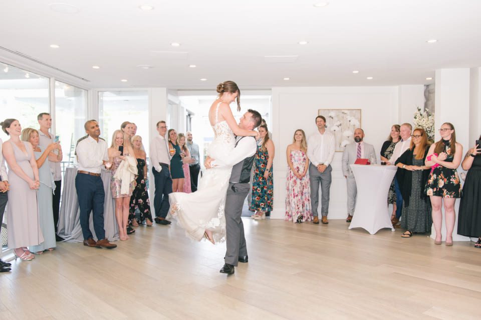A groom lifts the bride during their first dance in a bright room surrounded by guests. The bride wears a white gown, and the groom is in a gray suit. Guests watch, smiling and clapping, with some holding drinks and standing by cocktail tables. 124 on Queen Hotel and Spa | Hotel & Spa Niagara-on-the-lake | 1 (855) 988-4552