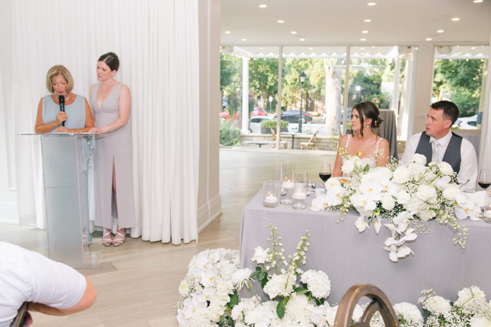 A woman speaks into a microphone next to another woman in a grey dress at a podium. A couple is seated at a table adorned with white flowers and candles, listening. Large windows reveal trees and parked cars outside. 124 on Queen Hotel and Spa | Hotel & Spa Niagara-on-the-lake | 1 (855) 988-4552
