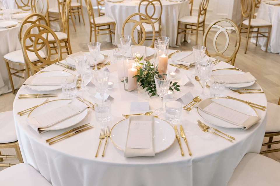 Elegant dining table set for an event, adorned with white plates, gold cutlery, crystal glasses, and cream napkins. Three pink candles and a small greenery centerpiece decorate the table, surrounded by gold chairs with circular backrests. 124 on Queen Hotel and Spa | Hotel & Spa Niagara-on-the-lake | 1 (855) 988-4552