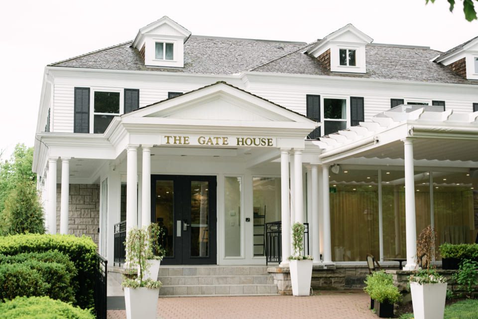 A two-story white building with dark shutters labeled The Gate House. It features a gabled roof, large windows, and a columned entrance. Green shrubs line the brick pathway leading to the entrance. 124 on Queen Hotel and Spa | Hotel & Spa Niagara-on-the-lake | 1 (855) 988-4552