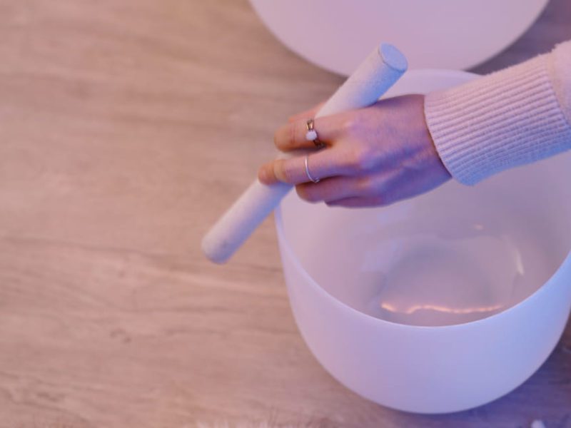 A person in a light sweater gently taps a frosted white singing bowl with a white mallet. They wear rings on their fingers. The scene is set on a wooden floor with a soft, serene tone. 124 on Queen Hotel and Spa | Hotel & Spa Niagara-on-the-lake | 1 (855) 988-4552
