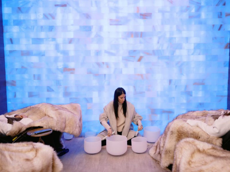A woman sits on the floor playing crystal singing bowls in a room with blue illuminated walls. Two people are reclining on chairs covered with fur blankets, facing her, creating a calm and serene atmosphere. 124 on Queen Hotel and Spa | Hotel & Spa Niagara-on-the-lake | 1 (855) 988-4552