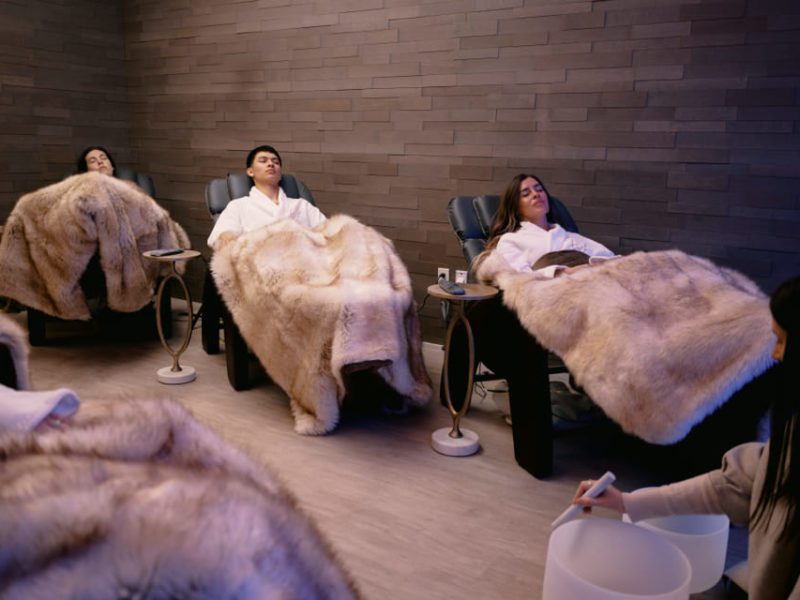Three individuals recline in cozy, modern chairs covered with faux fur blankets in a dimly lit room. A person at the forefront holds a singing bowl, suggesting a calming environment. 124 on Queen Hotel and Spa | Hotel & Spa Niagara-on-the-lake | 1 (855) 988-4552
