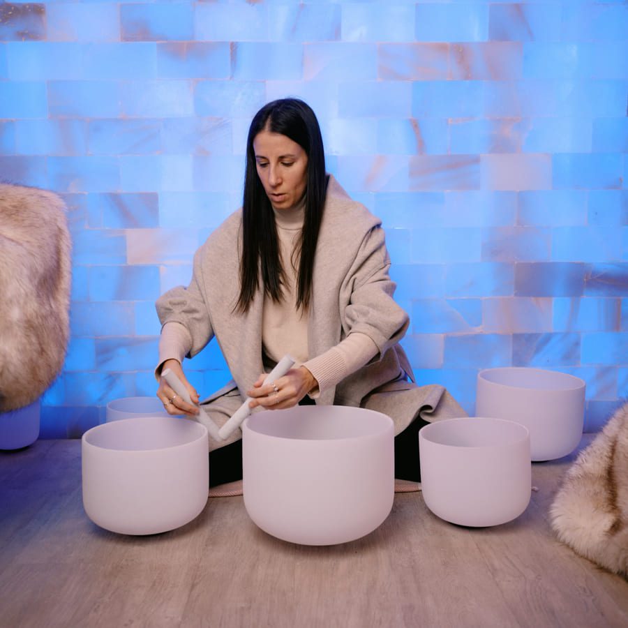 A person sitting cross-legged on the floor plays crystal singing bowls. They are in a room with a soothing blue-lit wall and faux fur blankets draped nearby. The scene exudes a calm and meditative atmosphere. 124 on Queen Hotel and Spa | Hotel & Spa Niagara-on-the-lake | 1 (855) 988-4552