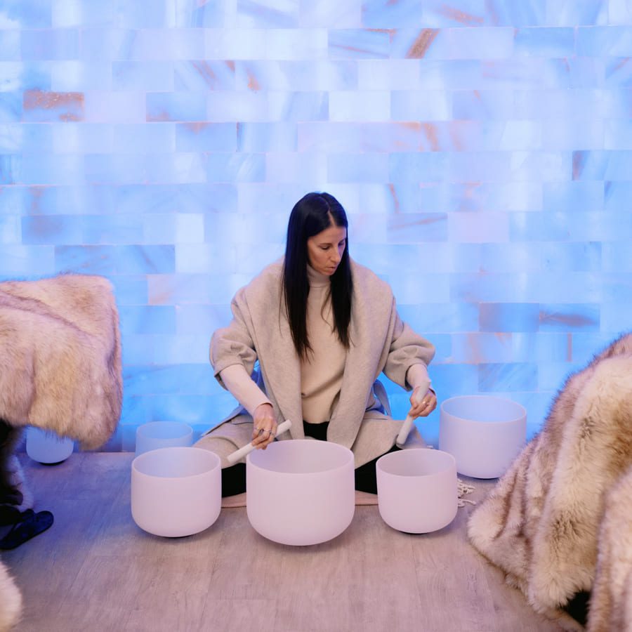 A person seated on the floor, playing white crystal singing bowls. They are wearing a cozy coat and are surrounded by fluffy blankets. The background features a light blue mosaic pattern. 124 on Queen Hotel and Spa | Hotel & Spa Niagara-on-the-lake | 1 (855) 988-4552