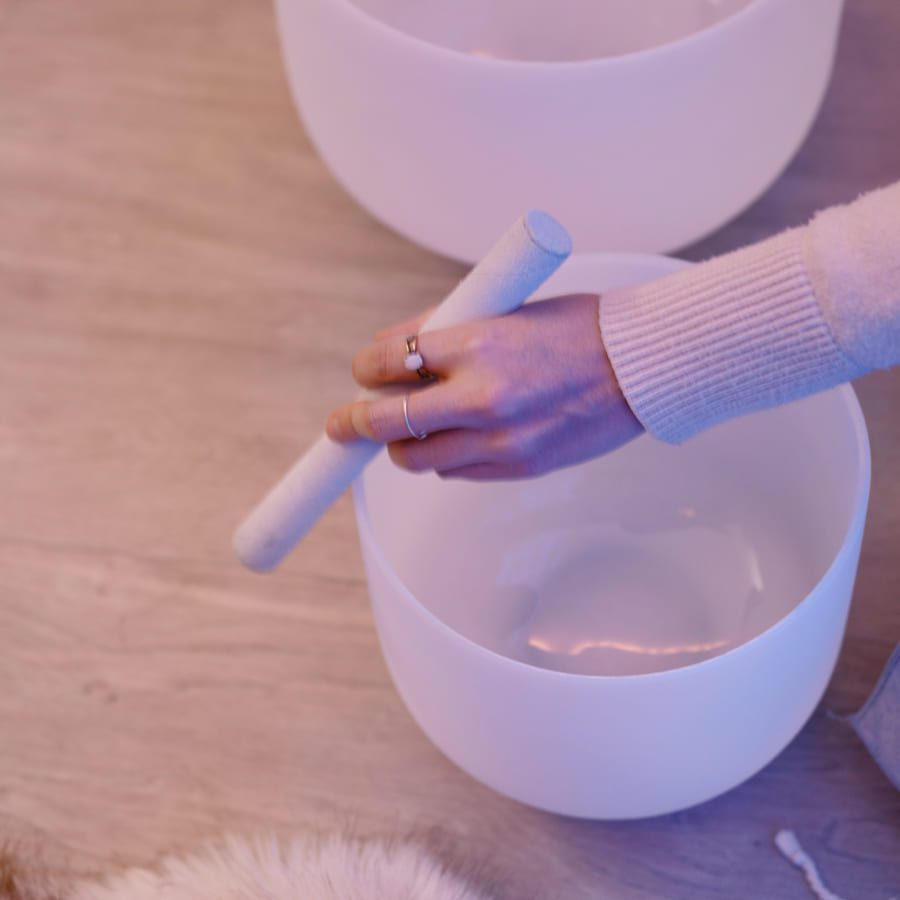 A hand holding a white mallet over a white singing bowl, resting on a light-colored wooden floor. The person is wearing a pale sweater and rings. A fuzzy, light-colored object is partially visible in the corner. 124 on Queen Hotel and Spa | Hotel & Spa Niagara-on-the-lake | 1 (855) 988-4552