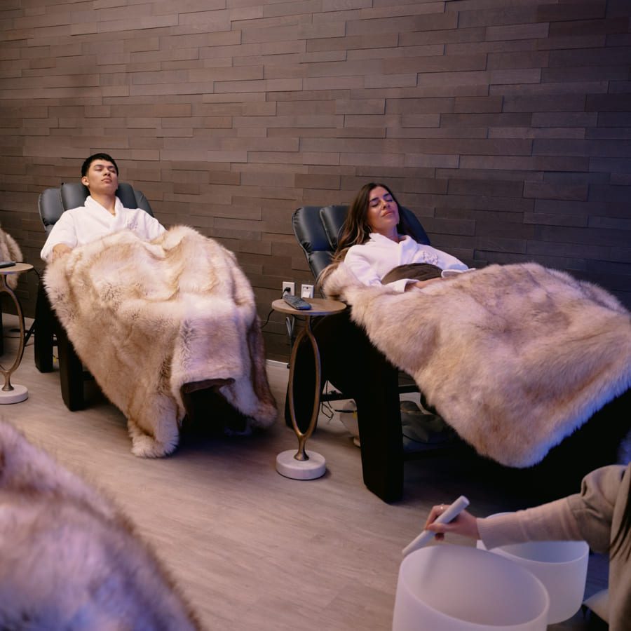 Two people relaxing in a spa environment. They are reclining in chairs, covered with plush faux fur blankets. The room has a warm, calming ambiance with soft lighting and a person in the foreground seated beside a small table. 124 on Queen Hotel and Spa | Hotel & Spa Niagara-on-the-lake | 1 (855) 988-4552