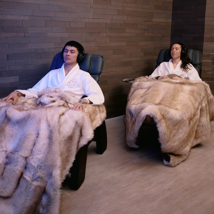 Two people are relaxing in reclined chairs, wearing white robes and large headphones. They are covered with plush fur blankets inside a room with dark brick walls. The atmosphere is calm and soothing. 124 on Queen Hotel and Spa | Hotel & Spa Niagara-on-the-lake | 1 (855) 988-4552