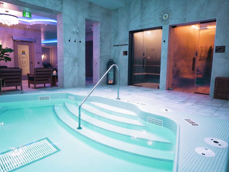 Indoor spa with a small pool featuring steps and water jets. The room has marble walls and neon lighting. Two steam rooms with glass doors are visible in the background, and there is a seating area with plants to the left. 124 on Queen Hotel and Spa | Hotel & Spa Niagara-on-the-lake | 1 (855) 988-4552