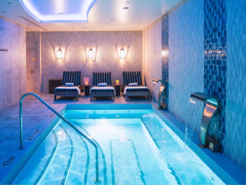 Luxurious indoor spa with a lit pool flanked by cascading water features. Three cushioned lounge chairs with striped towels are positioned against a patterned wall. Warm wall sconces and a soothing blue ambiance create a serene atmosphere. 124 on Queen Hotel and Spa | Hotel & Spa Niagara-on-the-lake | 1 (855) 988-4552