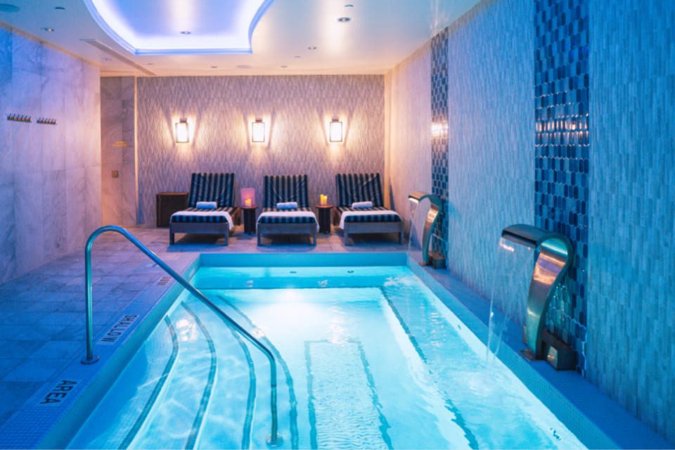 A luxurious indoor spa features a glowing blue pool with stainless steel massage jets. On the side, cushioned lounge chairs hold rolled towels, and the walls are elegantly tiled, illuminated by soft wall sconces. 124 on Queen Hotel and Spa | Hotel & Spa Niagara-on-the-lake | 1 (855) 988-4552