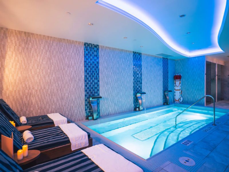 A luxurious indoor spa with a small glowing pool. Several cushioned lounge chairs with towels and a candle are set to the left. The ceiling features soft blue lighting, and the space is adorned with stylish tiles. 124 on Queen Hotel and Spa | Hotel & Spa Niagara-on-the-lake | 1 (855) 988-4552