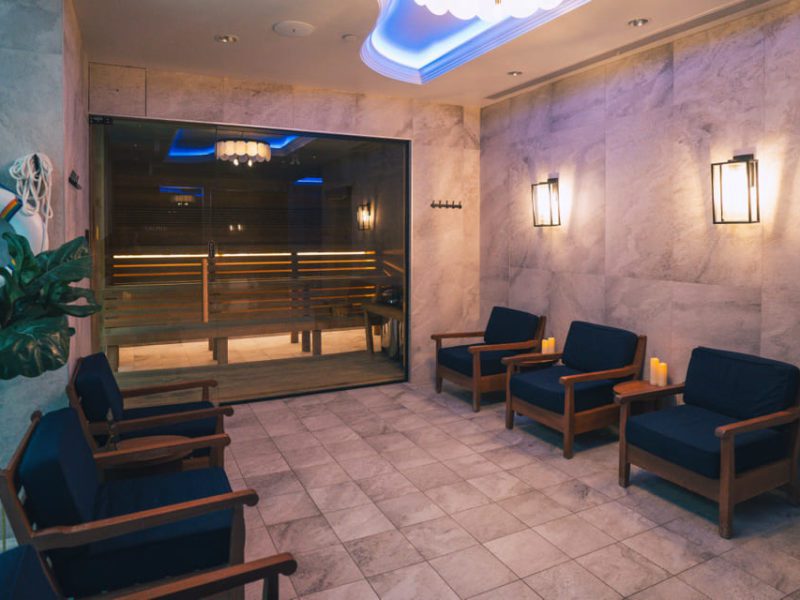 A cozy spa waiting area with several wooden chairs featuring navy cushions. Soft lighting from wall sconces and candles creates a warm ambiance. A large glass partition reveals a sauna inside, with visible wooden seating and soft lighting. 124 on Queen Hotel and Spa | Hotel & Spa Niagara-on-the-lake | 1 (855) 988-4552