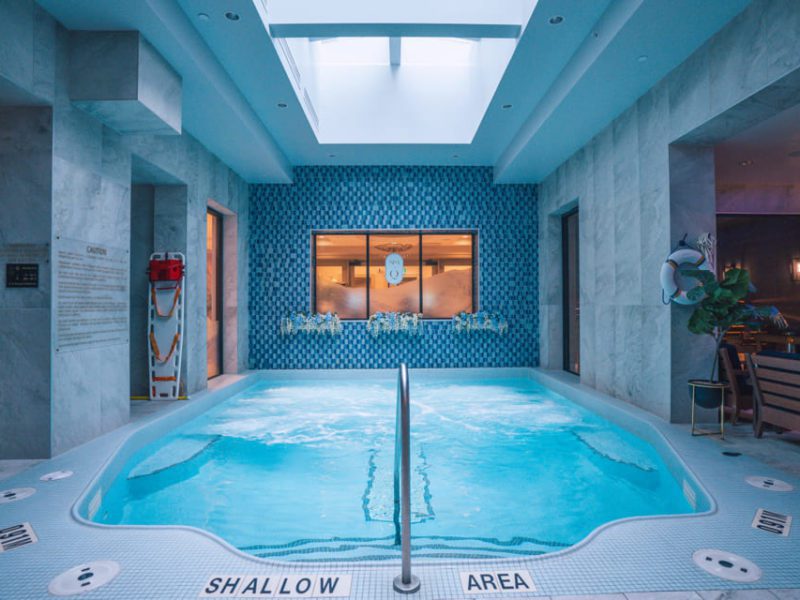 Indoor pool with light blue water, surrounded by a marble-like tiled deck. A skylight above illuminates the space. Shallow area is marked along the edge. Life-saving equipment is visible on the left, and a wall with a blue pattern is in the background. 124 on Queen Hotel and Spa | Hotel & Spa Niagara-on-the-lake | 1 (855) 988-4552