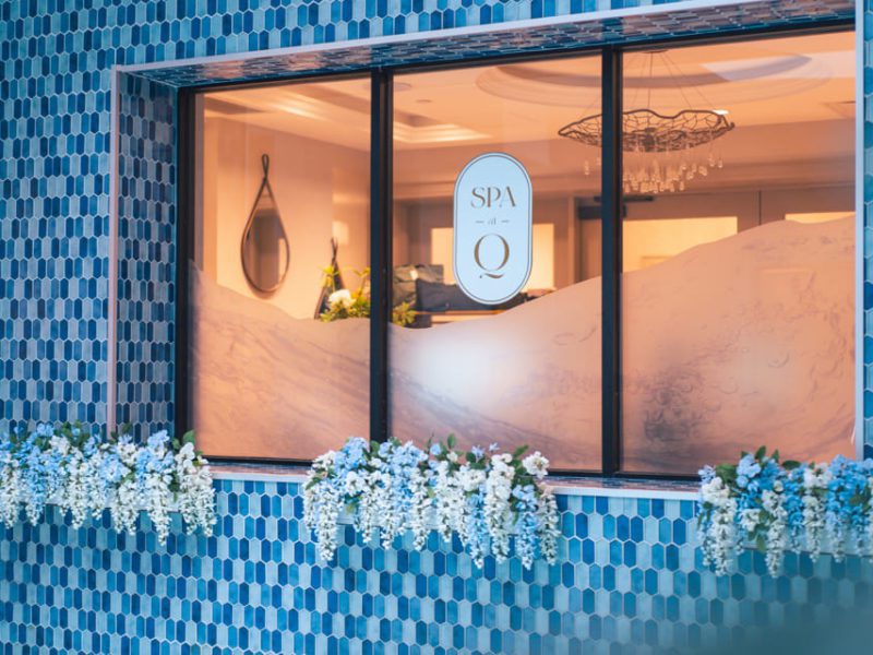 The image shows a window with a SPA logo, framed by blue patterned tiles. White flowers cascade from window boxes below. A chandelier and spa reception area are visible inside. 124 on Queen Hotel and Spa | Hotel & Spa Niagara-on-the-lake | 1 (855) 988-4552
