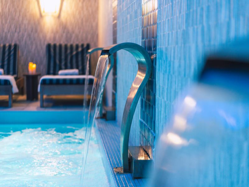 A luxurious indoor pool with cascading water features. Two cushioned lounge chairs with towels placed on them are in the background. The ambiance is serene with soft lighting and blue-tiled walls enhancing the tranquil atmosphere. 124 on Queen Hotel and Spa | Hotel & Spa Niagara-on-the-lake | 1 (855) 988-4552