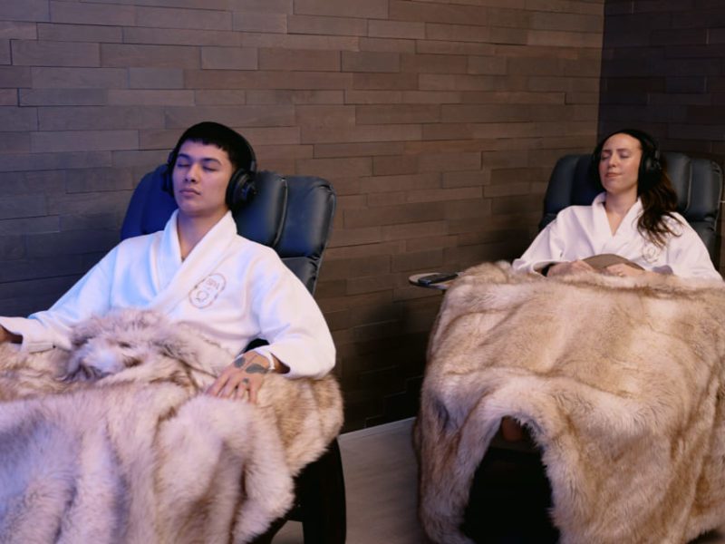 Two people relax in reclining chairs, each wearing white robes and headphones. They are covered with plush, fur-like blankets, sitting in a room with dark, textured walls, creating a cozy and calming atmosphere. 124 on Queen Hotel and Spa | Hotel & Spa Niagara-on-the-lake | 1 (855) 988-4552