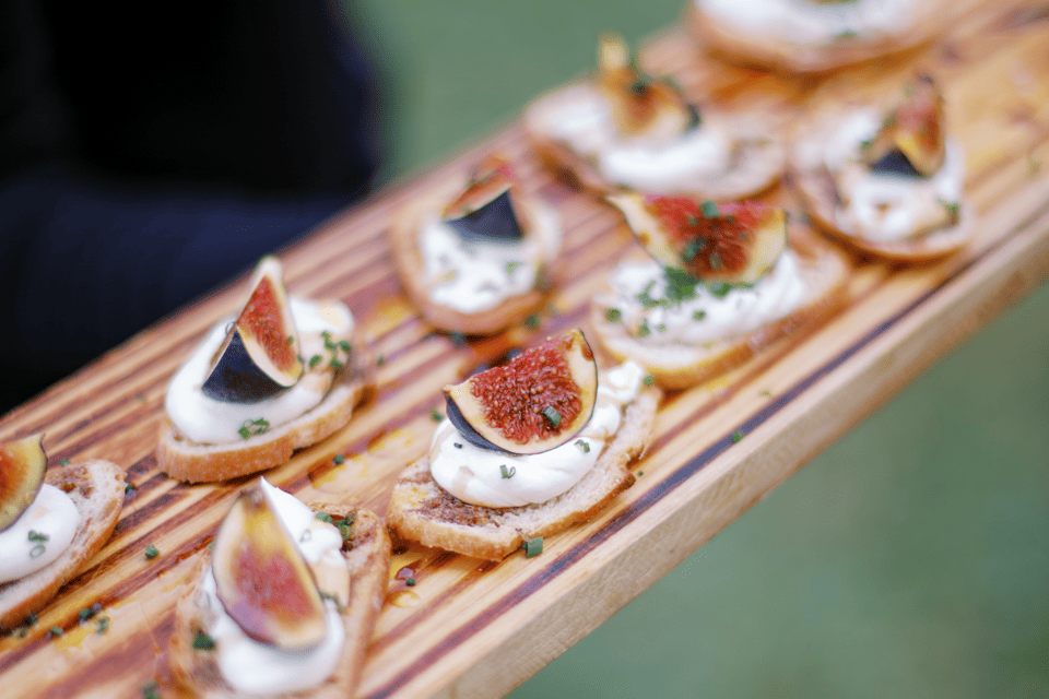 A wooden board displaying several slices of bread topped with a creamy spread, fresh fig slices, and sprinkled herbs. The background is softly blurred, highlighting the appetizers. 124 on Queen Hotel and Spa | Hotel & Spa Niagara-on-the-lake | 1 (855) 988-4552