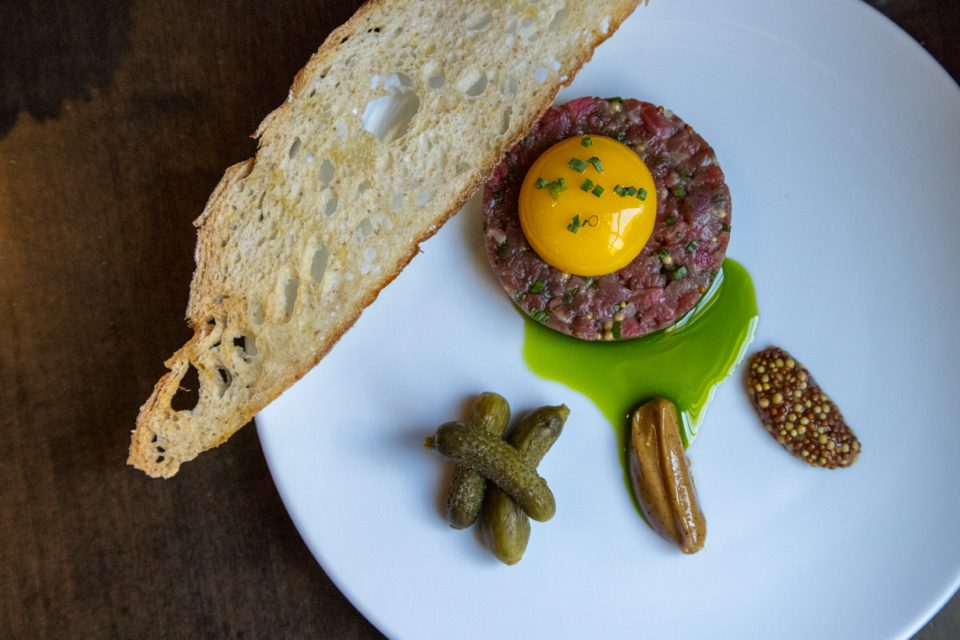 A beautifully arranged dish featuring beef tartare topped with a raw egg yolk and chives. Accompanied by a long slice of crusty bread, pickles, mustard, and a green sauce, all served on a white plate. 124 on Queen Hotel and Spa | Hotel & Spa Niagara-on-the-lake | 1 (855) 988-4552