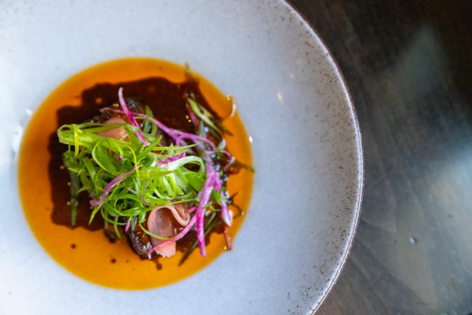 A round dish with braised meat in rich brown sauce, topped with green and purple shredded vegetables, served on a large white plate against a dark tabletop. 124 on Queen Hotel and Spa | Hotel & Spa Niagara-on-the-lake | 1 (855) 988-4552