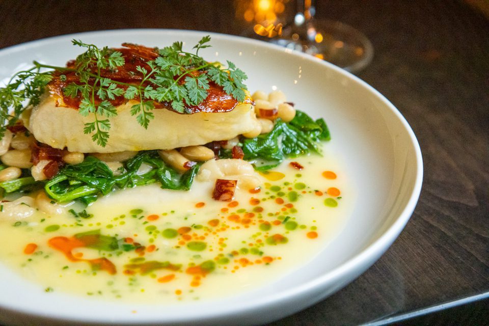 A beautifully plated dish featuring a seared piece of fish topped with fresh herbs, lying on a bed of sautéed spinach and white beans. The plate is garnished with colorful oil droplets and a rich sauce. 124 on Queen Hotel and Spa | Hotel & Spa Niagara-on-the-lake | 1 (855) 988-4552