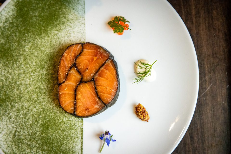 A gourmet dish with a circular arrangement of smoked salmon on a plate partially covered with green powder. Garnished with small herbs, a dot of sauce, mustard seeds, and a small flower. The plate is set against a dark wooden table. 124 on Queen Hotel and Spa | Hotel & Spa Niagara-on-the-lake | 1 (855) 988-4552