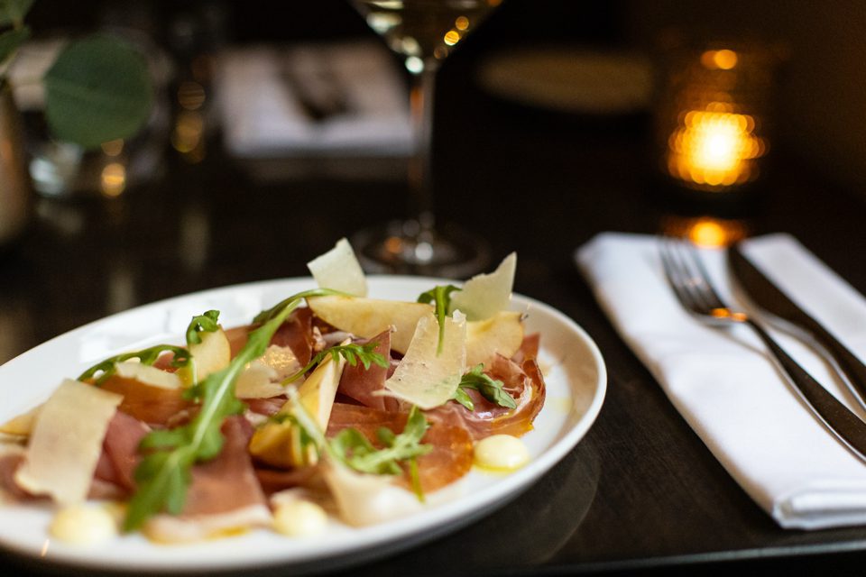A white plate on a dimly lit table holds thinly sliced prosciutto, shaved Parmesan, and arugula, with white sauce accents. A folded napkin with cutlery is beside it. A softly glowing candle and a wine glass are in the background. 124 on Queen Hotel and Spa | Hotel & Spa Niagara-on-the-lake | 1 (855) 988-4552
