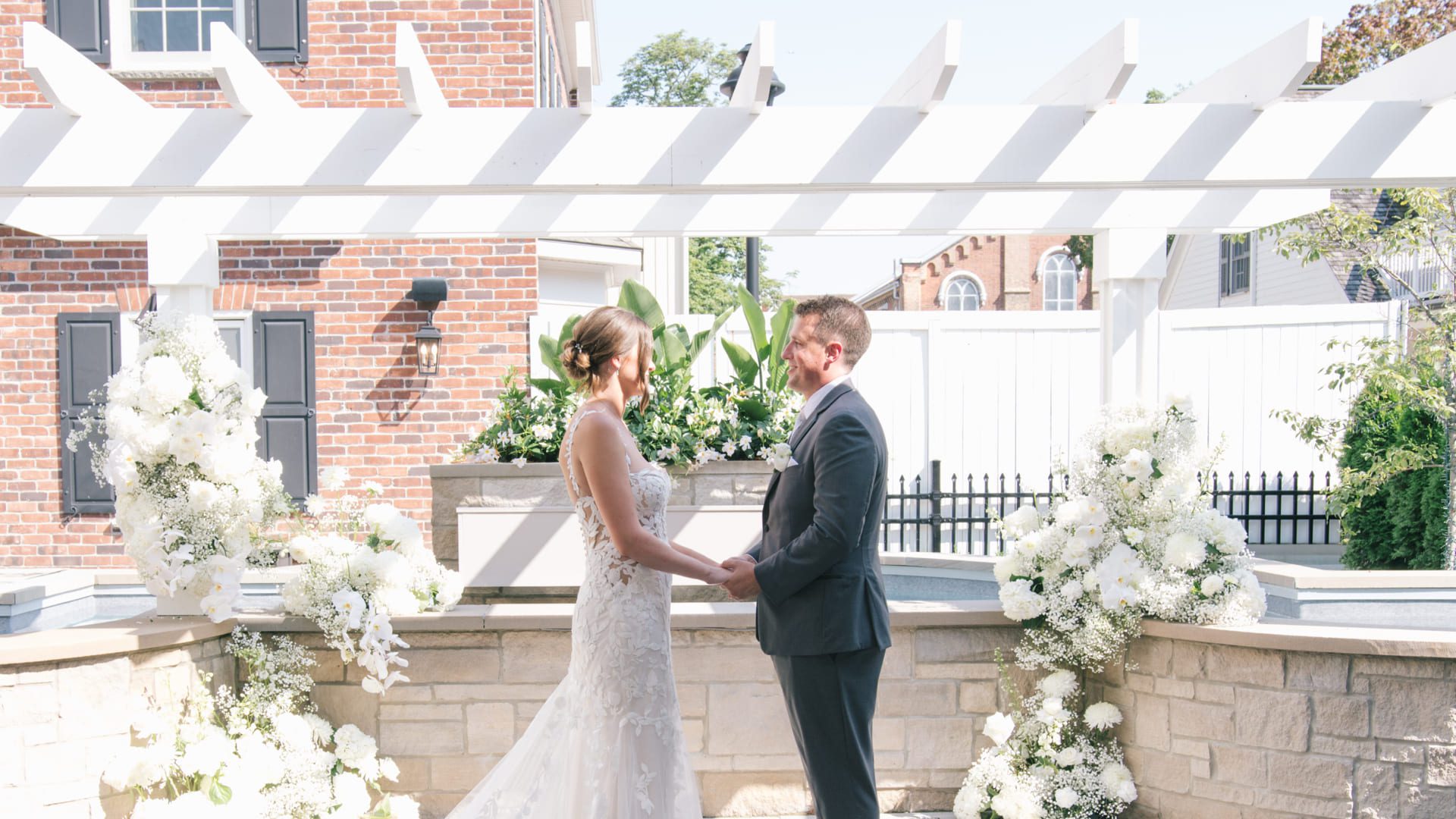A bride and groom hold hands, facing each other in an outdoor ceremony. The setting features white floral arrangements and a pergola, with a brick building in the background. The bride wears a lace gown, and the groom is in a dark suit. 124 on Queen Hotel and Spa | Hotel & Spa Niagara-on-the-lake | 1 (855) 988-4552