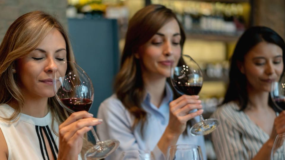 Three women are seated, each holding a glass of red wine, and they appear to be smelling the wine with eyes closed, suggesting they are tasting or evaluating it. Shelves with various bottles are visible in the blurred background. 124 on Queen Hotel and Spa | Hotel & Spa Niagara-on-the-lake | 1 (855) 988-4552